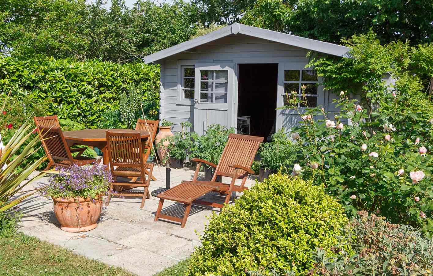 Gartenmöbel aus Holz vor einem Gartenhaus mit Fenstern und Spitzdach 